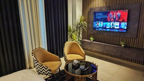 A modern hotel room with a large flat-screen TV displaying a movie, two armchairs, a small round coffee table, and a potted plant. - Penta Square - Luxury Studio Suite (Lahore)