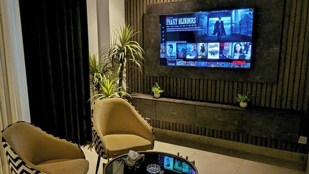 A modern living room with a flat-screen TV mounted on a wooden wall, a sofa, a coffee table with a screen, and a potted plant. - Penta Square - Luxury Studio Suite (Lahore)