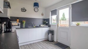 Et moderne køkken med hvide skabe, en sort bordplade, dekoration med kaffetema på væggen, et ur og et vindue med udsigt til et hegn og træer.
