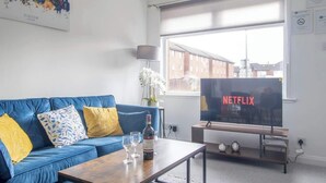 En moderne stue med en blå sofa, et sofabord med vinglas, et fjernsyn der viser 'NETFLIX' og et bybillede som kunst på væggen.