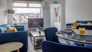 Et moderne hotelværelse med sofa, sofabord, et fjernsyn der viser 'NETFLIX', et lille bord med glas og en flaske, samt udsigt udad.