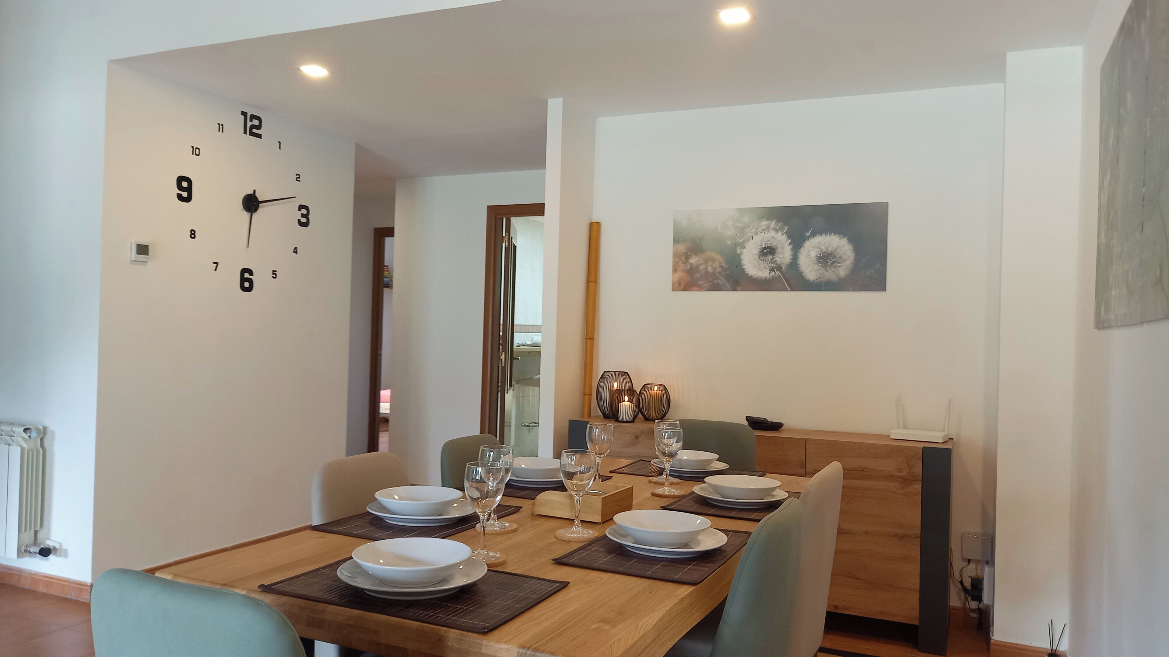 Uma sala de jantar com mesa de madeira posta para quatro pessoas, um relógio na parede e um quadro de dentes-de-leão.