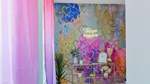 Um corredor com cortina rosa e roxa, um tapete estampado, parede com desenhos de corações, um letreiro neon e uma pequena mesa com objetos decorativos.