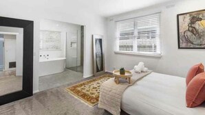 Een moderne slaapkamer met een groot bed, een klein rond tafeltje, een spiegel en een raam met jaloezieën.