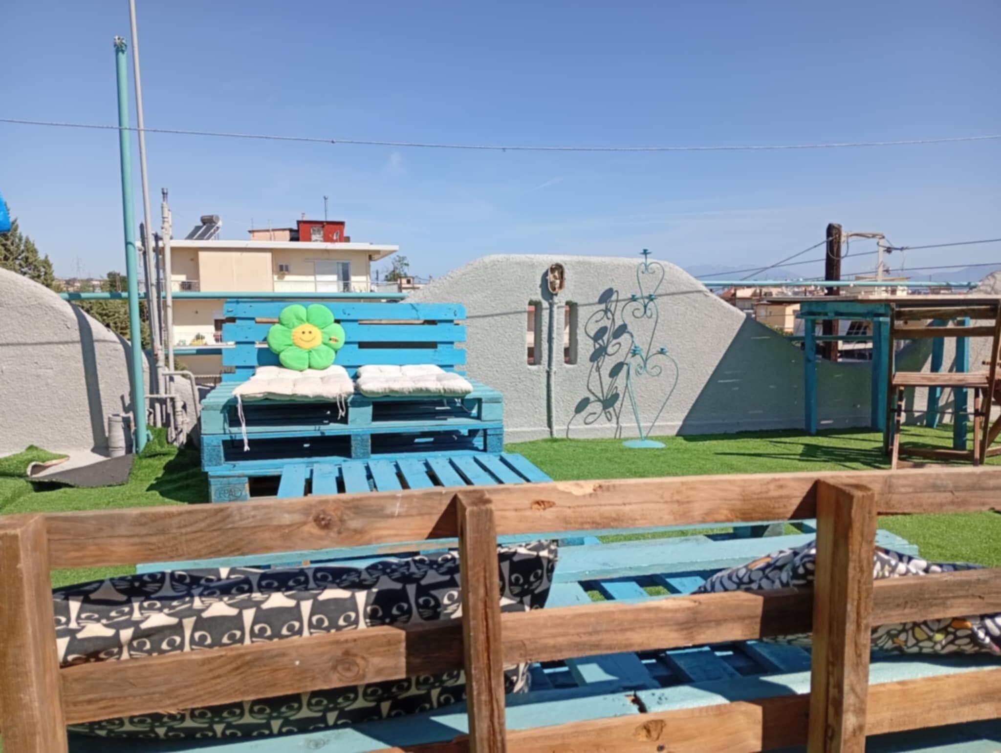 A wooden bench with patterned cushions, a blue bench with a flower cushion, and a white structure with a green roof.
