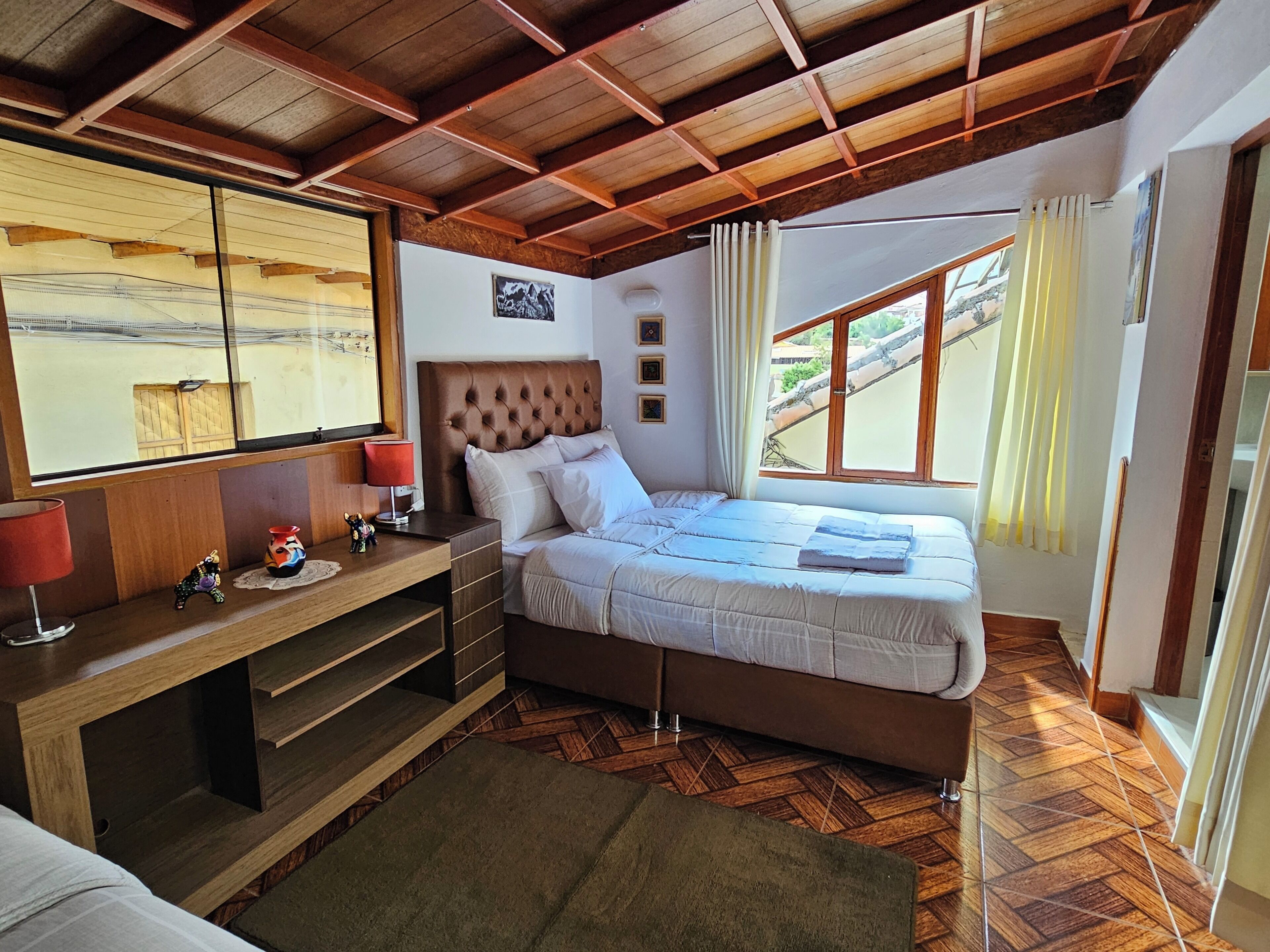 A bedroom with a wooden ceiling, a bed with white bedding, a nightstand with a lamp, and a window with curtains.