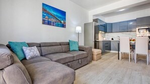 A modern living room with a grey sectional sofa, a painting of a cityscape, and a kitchen area with grey cabinets and white appliances. - The Apateu (San Pawl il-Baħar)