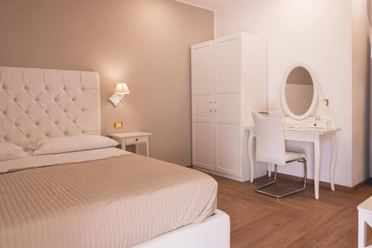 Un dormitorio con una cama de cabecera capitonada, una mesita de noche con lámpara, un armario, un escritorio con silla y un espejo redondo.