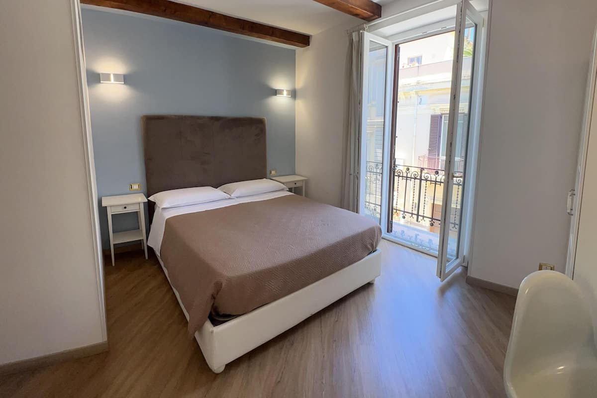 Un dormitorio con cama, mesitas de noche, una ventana con vistas al exterior y un balcón.