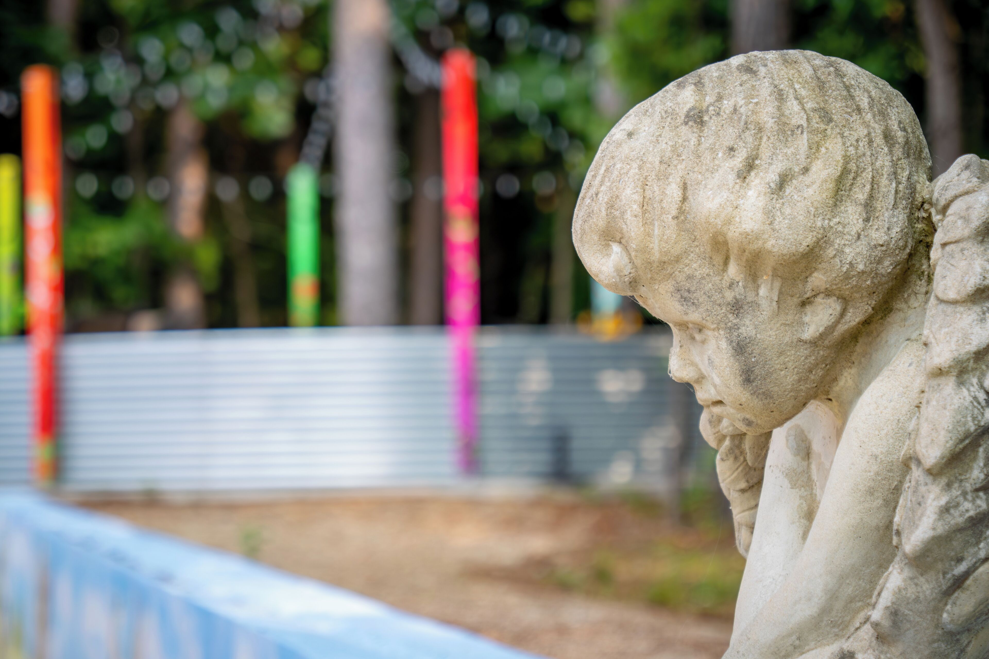A stone statue with a contemplative expression, set against a backdrop of trees and colorful poles.
