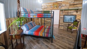 Un lit superposé avec une literie aux couleurs vives, une plante en macramé suspendue, une table en bois et un mur rustique orné de tableaux.