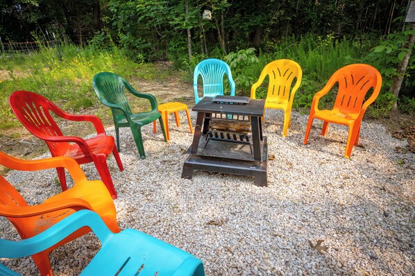 A group of colorful plastic chairs arranged around a small fire pit on a gravel surface, surrounded by greenery.