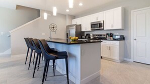 Una cocina moderna con armarios blancos, un refrigerador de acero inoxidable, un microondas y una estufa. Hay tres taburetes de bar con asientos y patas oscuras.