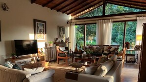 A spacious living room with a large sofa set, a television, and a coffee table. There are large windows providing a view of the outdoors. - Country House with Pool and Cinematic View in Luxury Condominium (Pedro do Rio)