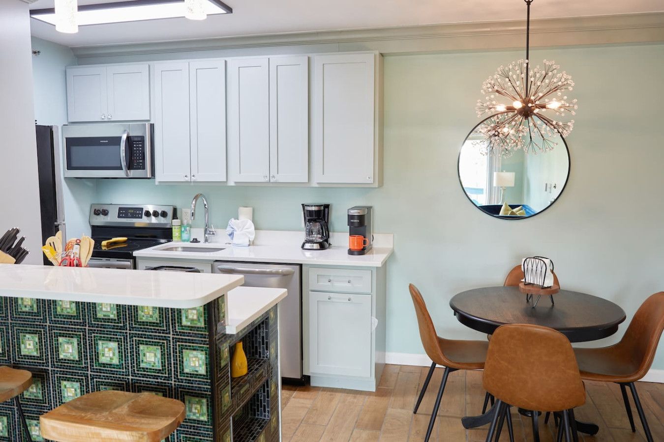 Una cocina moderna con armarios blancos, microondas, estufa y cafetera. Hay un comedor con una mesa redonda y sillas.