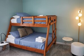 En køjeseng med træramme, blåt sengetøj og puder. To sengeborde, hvoraf det ene har en lampe og det andet et rundt spejl. En gulvlampe med flere lyskilder er placeret på højre side.