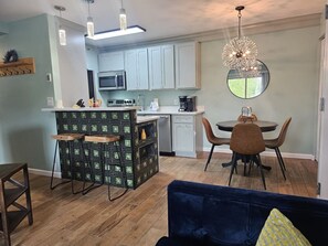 A modern kitchen with light blue cabinets, a central island with stools, a dining area with a round table and chairs, and a large mirror above the dining table.