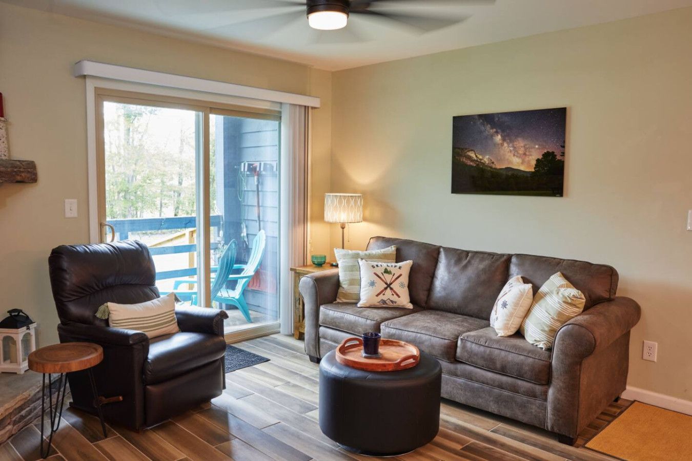 A living room with a leather sofa, a leather armchair, a wooden side table, and a round ottoman. There is a sliding glass door leading to an outdoor area with a blue chair and a table. A painting of a night sky is on the wall.