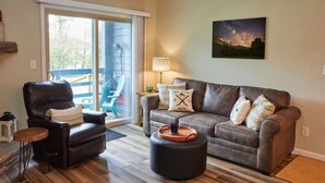 Una sala de estar con un sofá de cuero, un sillón de cuero, una mesita de madera y un reposapiés redondo. Hay una puerta corrediza de vidrio que da a un área exterior con una silla azul y una mesa. Un cuadro de un cielo nocturno está en la pared.
