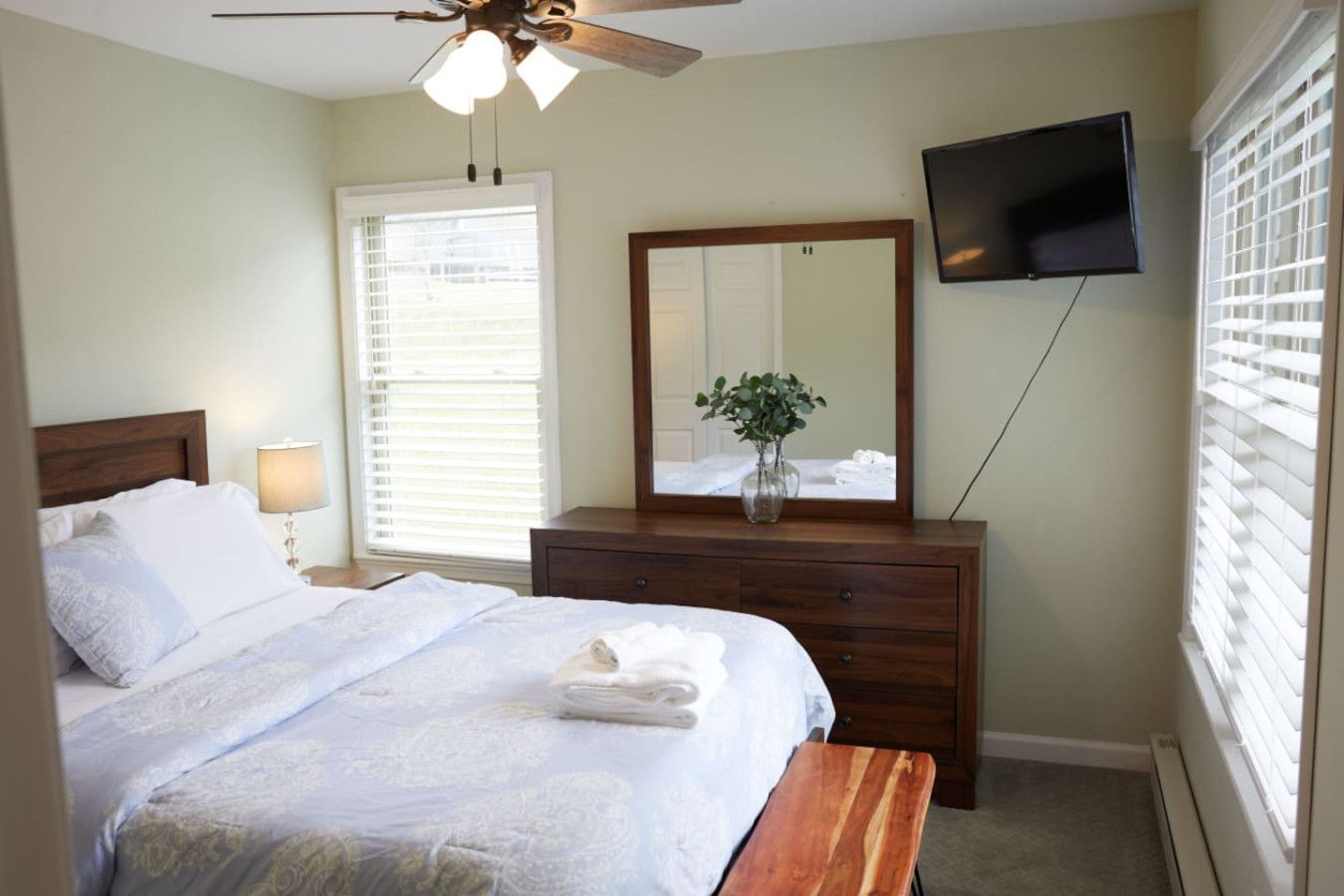 A bedroom with a bed, bedside table, lamp, dresser, mirror, TV, and window with blinds.