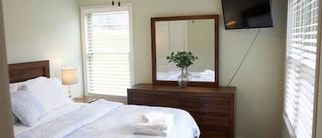 A bedroom with a bed, bedside table, lamp, dresser, mirror, TV, and window with blinds.