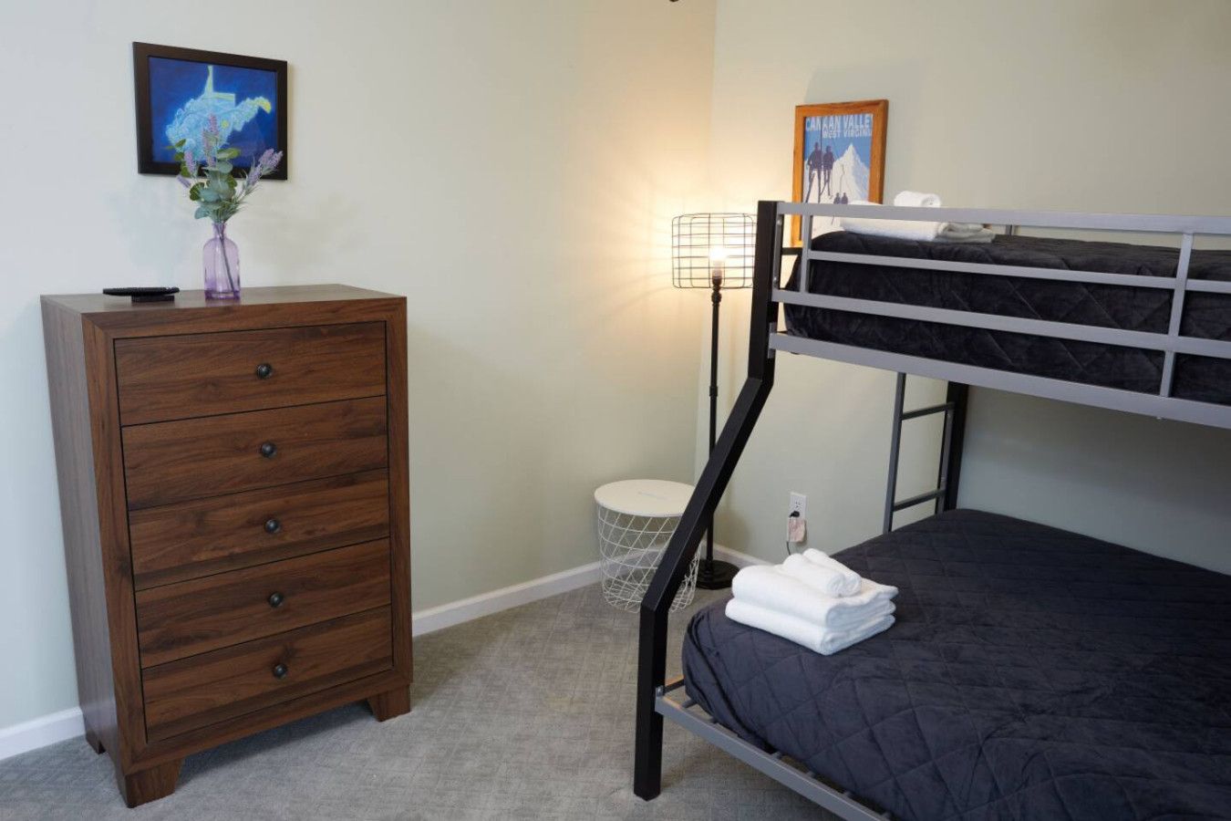 A bunk bed with a dark quilted mattress, a wooden dresser with four drawers, a small round table with a wireframe lamp, and a framed picture on the wall.
