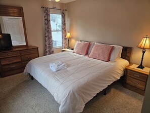 Een netjes opgemaakt bed met een witte deken en rode kussens, een houten nachtkastje met een lamp, een houten dressoir en een raam met gordijnen.