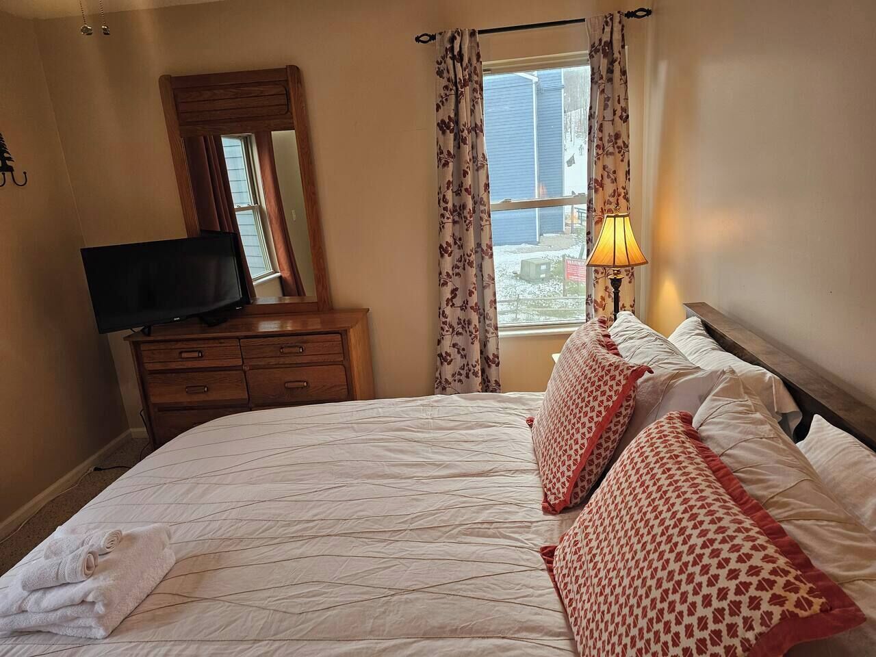 A neatly made bed with patterned pillows, a bedside lamp, a dresser with a flat-screen TV, and a window with curtains.