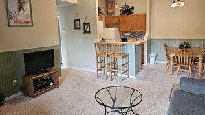 A living room with a television, a dining area with chairs and a table, and a kitchen area with wooden cabinets and a refrigerator.