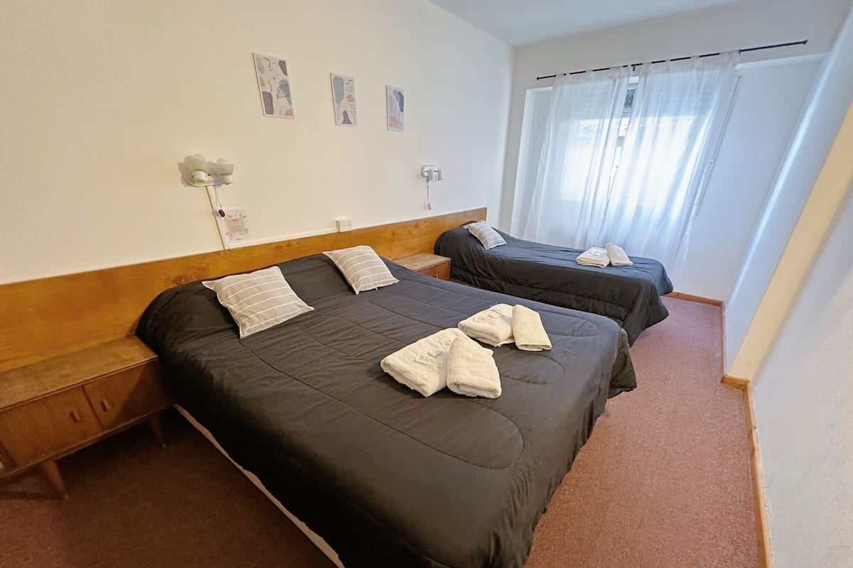 Une chambre d’hôtel avec deux lits, une tête de lit en bois, une petite table de chevet et une fenêtre avec des rideaux légers.
