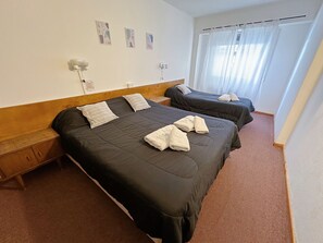 Habitación de hotel con dos camas, cabecero de madera, mesita de noche pequeña y ventana con cortinas de gasa.