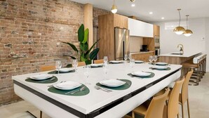 Un comedor moderno con pared de ladrillo, una mesa larga dispuesta para seis comensales y una cocina al fondo.