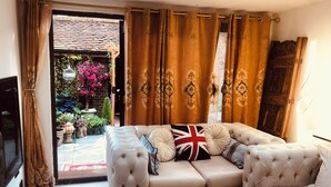 A living room with a beige sofa, a round coffee table, and a view of a garden with pink flowers and a brick wall. - London Central 4Bed House with Garden (London)
