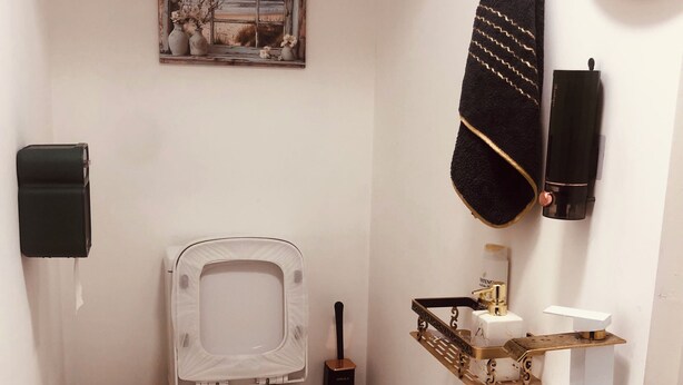 Un baño pequeño con inodoro, lavamanos con grifo dorado, un cubo de basura y una toalla decorativa.
