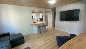 A modern living room with a wooden ceiling, a flat-screen TV mounted on the wall, and a kitchen area with white cabinets and a bar stool. - Cozy cabin with WiFi in charming Au Train, Completely remodeled as of June 2025 (Au Train)