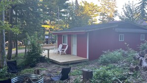A red cabin with a deck, two white chairs, a metal fire pit, and surrounding forest. - Cozy cabin with WiFi in charming Au Train, Completely remodeled as of June 2025 (Au Train)