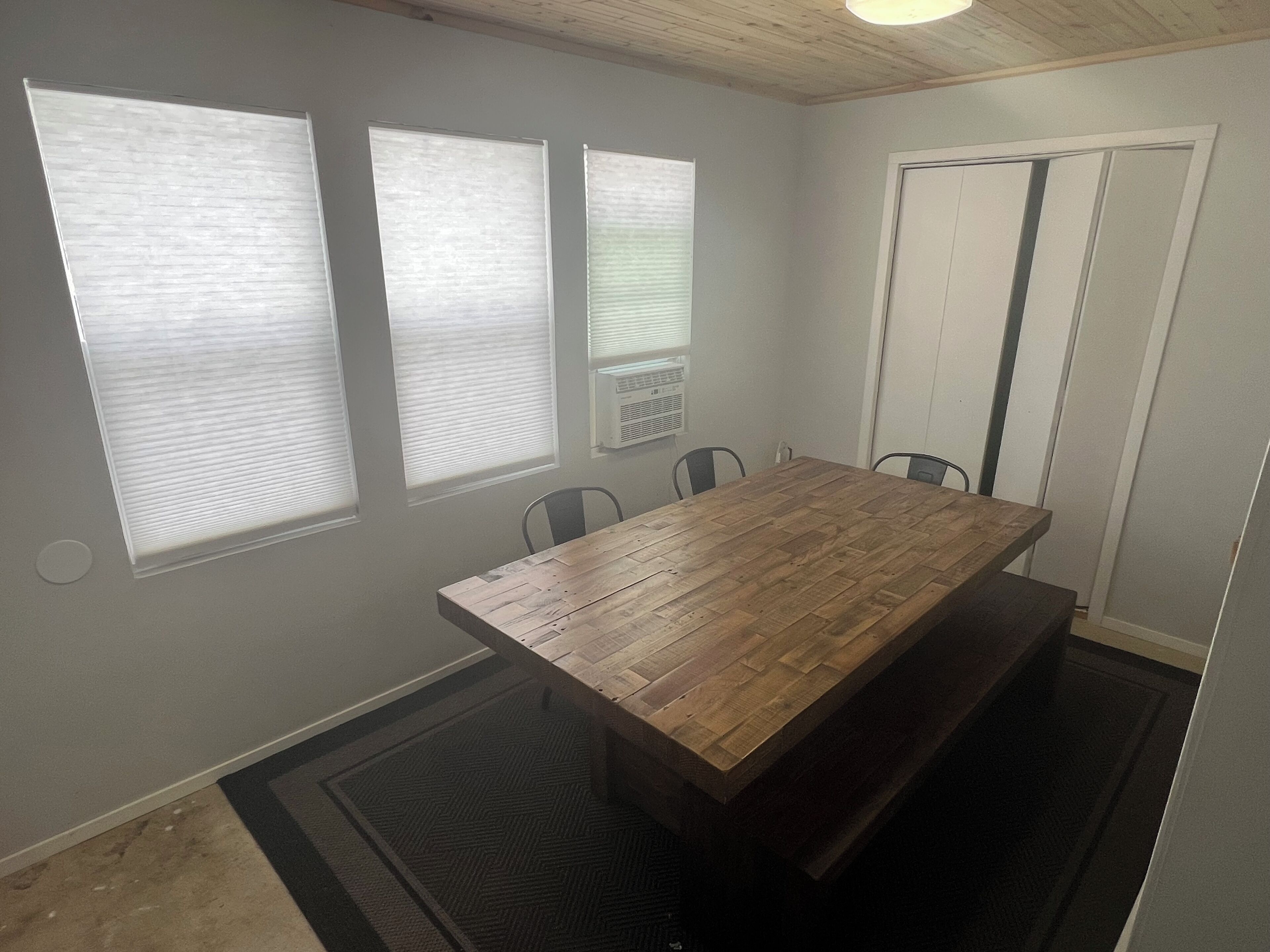 Une pièce avec une table en bois, trois fenêtres équipées de stores, une porte et un sol recouvert d’une moquette.