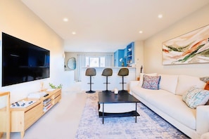 A modern living room with a flat-screen TV, a sofa, a coffee table, and a kitchen area in the background.