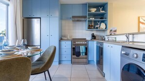 A modern kitchen with blue cabinets, a dining area with a glass table, and a built-in washing machine. - Casino Margaret Mahy  2bd garage (Christchurch)