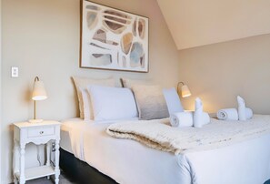 A neatly made bed with white linens and a fluffy white throw blanket, a bedside table with a lamp, and a wall-mounted abstract art piece.