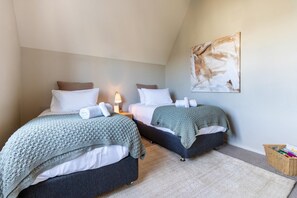 Une chambre double avec deux lits, chacun recouvert d’une couverture tricotée grise et agrémenté d’oreillers blancs. On y trouve une table de chevet avec une lampe, une œuvre d’art accrochée au mur et un panier en osier.