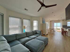 A spacious living area with a large grey sectional sofa, a ceiling fan, and a view of the ocean through the sliding glass doors.