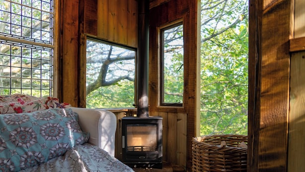 A cozy cabin interior with a bed, a wood-burning stove, and large windows overlooking a forest. - Treehouse Cabin Wilderness Escape (Abergele)