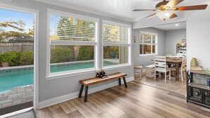 A room with a wooden bench, a dining table with chairs, a ceiling fan, and large windows overlooking a swimming pool.