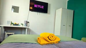 Ein Hotelzimmer mit einem Bett, einem Schreibtisch mit Computer, einem Kleiderschrank und einem Fernseher, der 'NETFLIX' anzeigt.