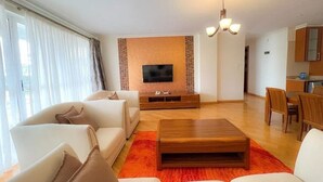 A modern living room with a flat-screen TV, a wooden coffee table, and a dining area with wooden chairs. - Luxurious & Exclusively furnished Suite -UN Blue Zone, Child Friendly Apt. (Nairobi)