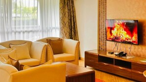A living room with a sofa set, a flat-screen TV displaying a fire, and a wooden entertainment unit. - Luxurious & Exclusively furnished Suite -UN Blue Zone, Child Friendly Apt. (Nairobi)