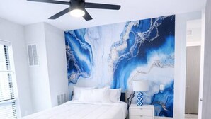 A bedroom with a bed, a nightstand, a lamp, a ceiling fan, and a large abstract wall mural. - Lightning Luxe Retreat (Des Moines)