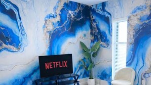 A living room with a blue sofa, a coffee table, a TV, and a large abstract mural on the wall. - Lightning Luxe Retreat (Des Moines)