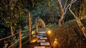 A stone pathway leading through a forested area with string lights and a wooden structure in the distance.
